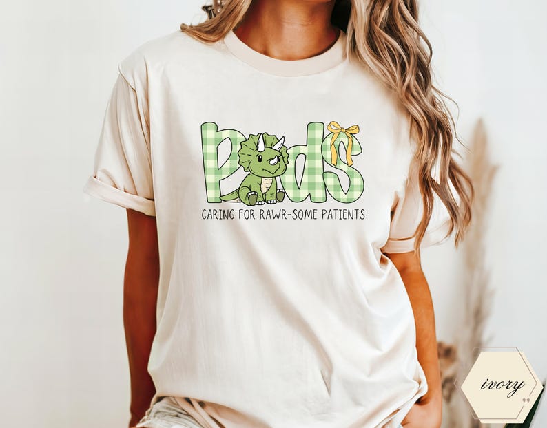 May include: Ivory t-shirt with the word "Peds" in green plaid letters and a cartoon dinosaur. The shirt also features the phrase "Caring for Rawr-some Patients" in black text. A yellow bow accents the "ds" in "Peds".