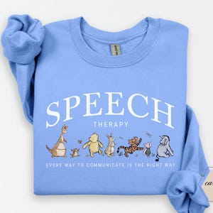 May include: A light blue sweatshirt with the words "SPEECH THERAPY" in white arched text. Below the text is a line of cartoon characters. The phrase "EVERY WAY TO COMMUNICATE IS THE RIGHT WAY" is printed below the characters.