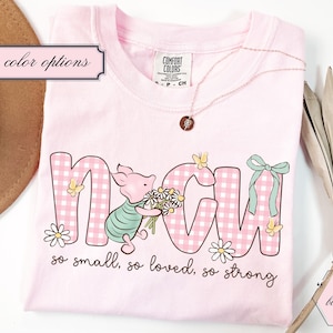 May include: A light pink t-shirt with the word "MOM" in a pink gingham pattern, featuring a pig holding flowers. The shirt includes the text "so small, so loved, so strong". A gold necklace and a decorative leaf are also visible.