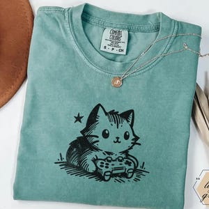 May include: Light green t-shirt featuring a black graphic of a cat playing a video game. The shirt has a crew neck and short sleeves. A gold necklace with a pendant is draped across the shirt. The tag reads "Comfort Colors".
