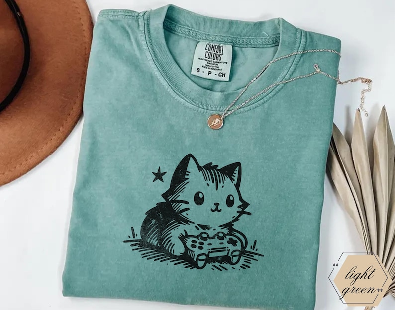 May include: Light green t-shirt featuring a black graphic of a cat playing a video game. The shirt has a crew neck and short sleeves. A gold necklace with a pendant is draped across the shirt. The tag reads "Comfort Colors".