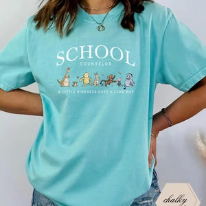 May include: A mint green short-sleeve t-shirt featuring the white text SCHOOL COUNSELOR above a line of illustrated characters including Kanga, Roo, Winnie the Pooh, Rabbit, Tigger, Piglet, and Eeyore, with the text A LITTLE KINDNESS GOES A LONG WAY.