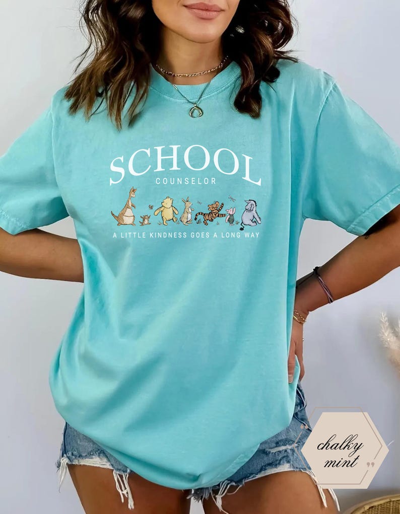 May include: A mint green short-sleeve t-shirt featuring the white text SCHOOL COUNSELOR above a line of illustrated characters including Kanga, Roo, Winnie the Pooh, Rabbit, Tigger, Piglet, and Eeyore, with the text A LITTLE KINDNESS GOES A LONG WAY.