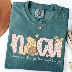 May include: Teal Comfort Colors t-shirt with the word "NCU" in a pink heart pattern, a Winnie the Pooh graphic, and the phrase "a hug is always the right size." A gold necklace and a brown hat are also visible.