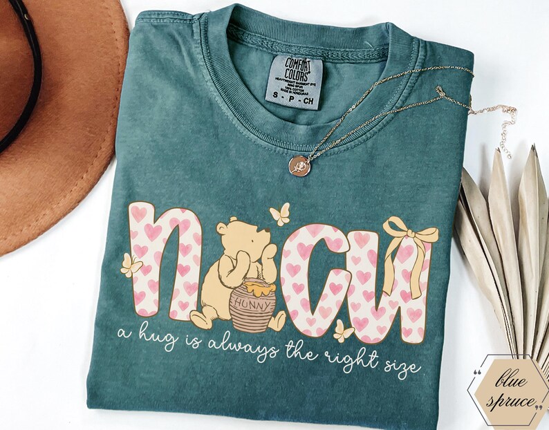 May include: Teal Comfort Colors t-shirt with the word "NCU" in a pink heart pattern, a Winnie the Pooh graphic, and the phrase "a hug is always the right size." A gold necklace and a brown hat are also visible.