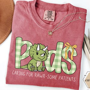 May include: A red t-shirt featuring the word "Peds" in green plaid lettering and a cartoon triceratops. The text "Caring for Rawr-some Patients" is below the dinosaur. A gold necklace is draped across the shirt.
