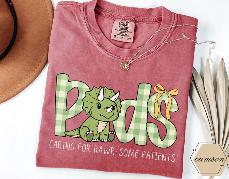 May include: A red t-shirt featuring the word "Peds" in green plaid lettering and a cartoon triceratops. The text "Caring for Rawr-some Patients" is below the dinosaur. A gold necklace is draped across the shirt.