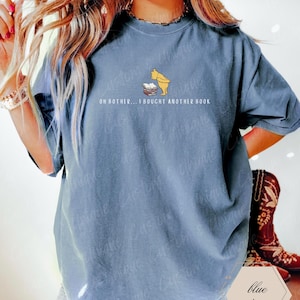 May include: Blue jean colored t-shirt featuring a cartoon illustration of Winnie the Pooh looking at a stack of books. The text "OH BOTHER... I BOUGHT ANOTHER BOOK" is printed below the illustration. The shirt has short sleeves.