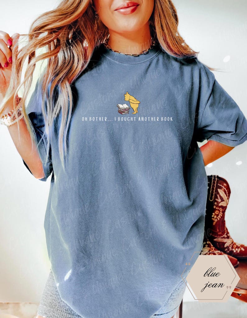 May include: Blue jean colored t-shirt featuring a cartoon illustration of Winnie the Pooh looking at a stack of books. The text "OH BOTHER... I BOUGHT ANOTHER BOOK" is printed below the illustration. The shirt has short sleeves.