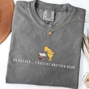 May include: A gray Comfort Colors t-shirt featuring a graphic of Winnie the Pooh with books and the text "OH BOTHER... I BOUGHT ANOTHER BOOK." The shirt has a crew neck and short sleeves. A gold necklace is draped across the shirt.