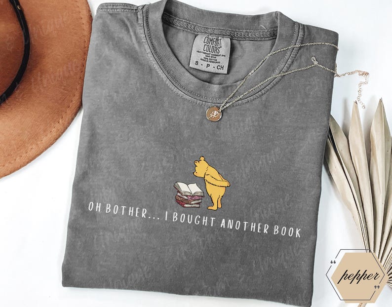 May include: A gray Comfort Colors t-shirt featuring a graphic of Winnie the Pooh with books and the text "OH BOTHER... I BOUGHT ANOTHER BOOK." The shirt has a crew neck and short sleeves. A gold necklace is draped across the shirt.