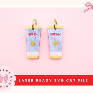 May include: A pair of earrings shaped like sunscreen tubes. Each earring has a gold-colored hoop, a white base, and a blue and white tube with the text "SPF 50+" and a yellow sun graphic. The background is pink.