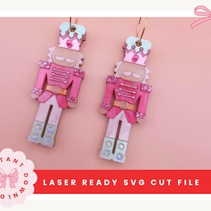 Könnte beinhalten: Zwei rosa und glitzernde Nussknacker-Ohrringe mit goldenen Reifen. Die Ohrringe haben eine rosa Uniform, eine Krone und einen Schnurrbart. Die Ohrringe sind vor einem rosa Hintergrund platziert. Der Text "LASER READY SVG CUT FILE" ist sichtbar.