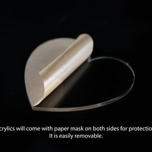 May include: A clear acrylic heart shape with a brown paper protective mask on one side. The mask is partially peeled back to reveal the clear acrylic.