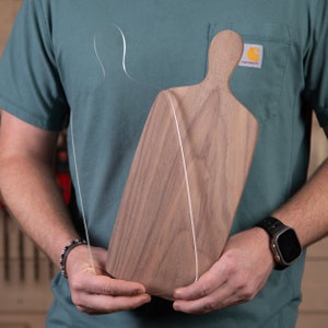 May include: A wooden cutting board with a handle is held in front of a person. The board is made of dark brown wood and has a smooth surface. A clear acrylic outline of a person is attached to the board with a white string.