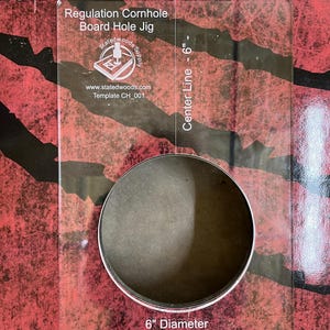 May include: A clear acrylic regulation cornhole board hole jig. The jig has a 6-inch diameter hole and is marked with a center line. Text on the jig includes "Regulation Cornhole Board Hole Jig" and "Statedwoods Supply".
