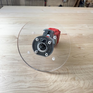 May include: A red and black power router with a clear circular base, resting on a light-colored wooden surface. The tool is designed for woodworking and is positioned in the center of the frame.