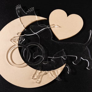 May include: A wooden crescent moon cutout with a heart and a ring with a diamond, all on a black background. There are also two clear acrylic cutouts of dogs, one with a tail and one without.