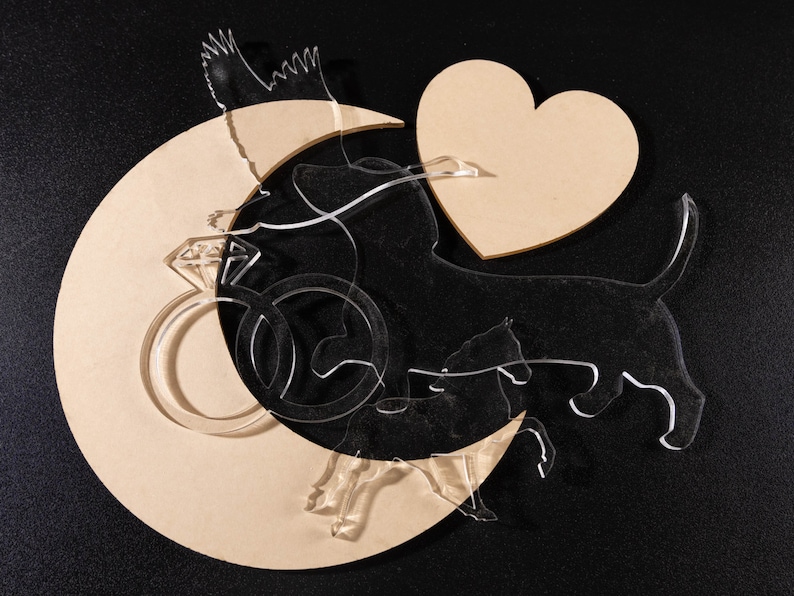 May include: A wooden crescent moon cutout with a heart and a ring with a diamond, all on a black background. There are also two clear acrylic cutouts of dogs, one with a tail and one without.