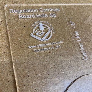 May include: Clear acrylic cornhole board hole jig with the text "Regulation Cornhole Board Hole Jig." The jig includes a logo and the text "Center Line - 6". The template is for a 6-inch hole.