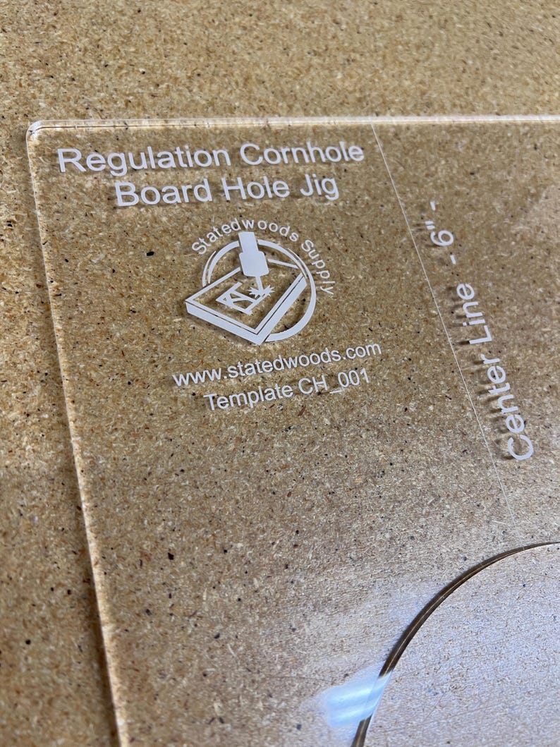 May include: Clear acrylic cornhole board hole jig with the text "Regulation Cornhole Board Hole Jig." The jig includes a logo and the text "Center Line - 6". The template is for a 6-inch hole.