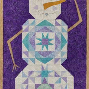 May include: A quilted snowman wall hanging. The snowman has a tan hat, an orange carrot nose, and a smiling face. The body is made of white, blue, and purple fabric. The background is purple with white snowflake patterns.