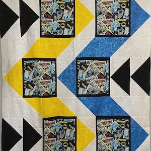May include: A white, yellow, blue, and black quilt with a geometric pattern. The quilt features a repeating pattern of squares with a comic book design featuring the word "Batman".