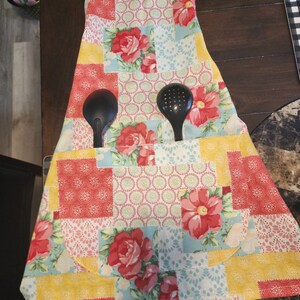 May include: A patchwork kitchen apron with a floral design in shades of pink, red, yellow, and blue. The apron has a large pocket in the front and two black kitchen utensils are tucked into the pocket.