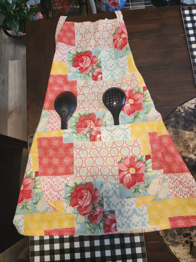 May include: A patchwork kitchen apron with a floral design in shades of pink, red, yellow, and blue. The apron has a large pocket in the front and two black kitchen utensils are tucked into the pocket.