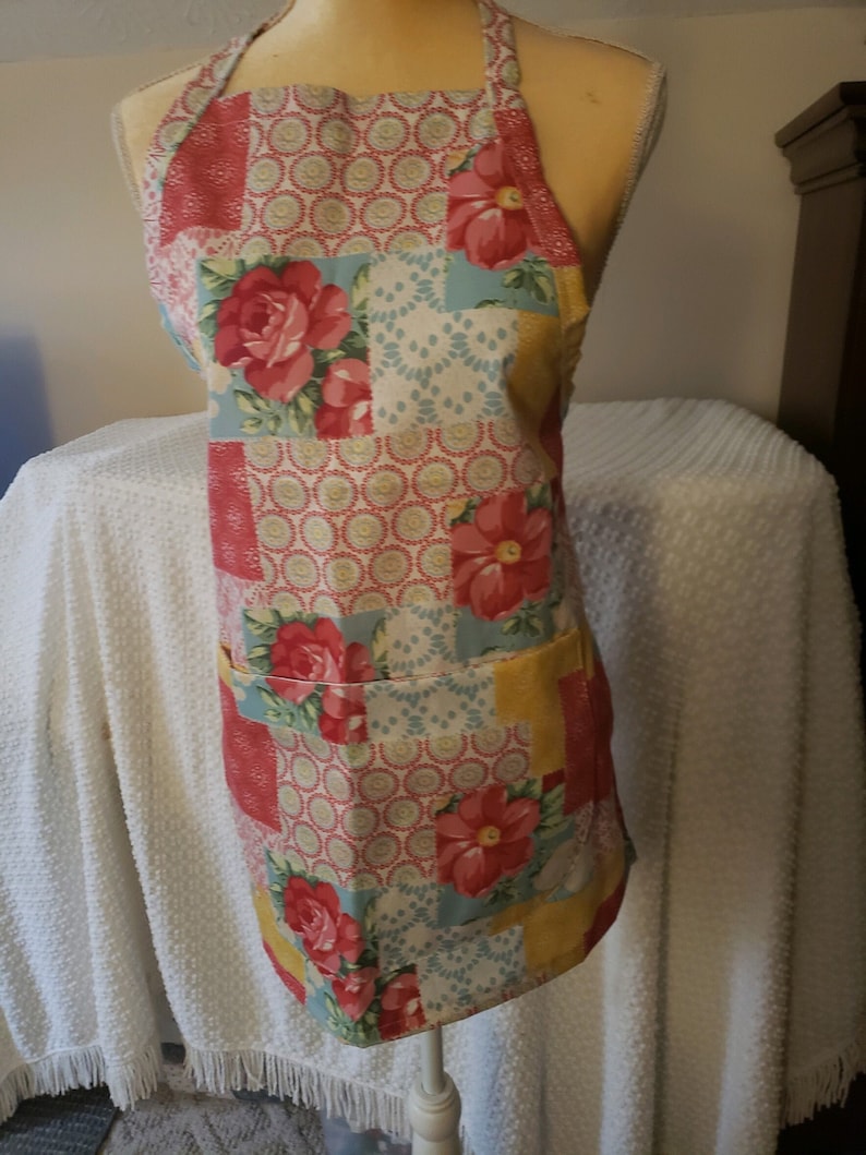May include: A pink, blue and yellow patchwork apron with a floral pattern. The apron has a pocket on the front.