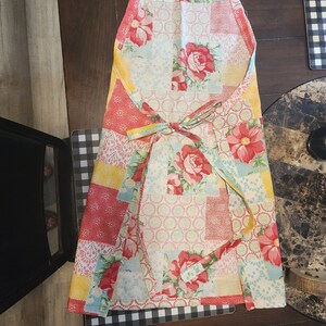 May include: A colourful patchwork apron with a floral pattern in shades of red, yellow, blue, and white. The apron has adjustable straps and a tie at the waist.