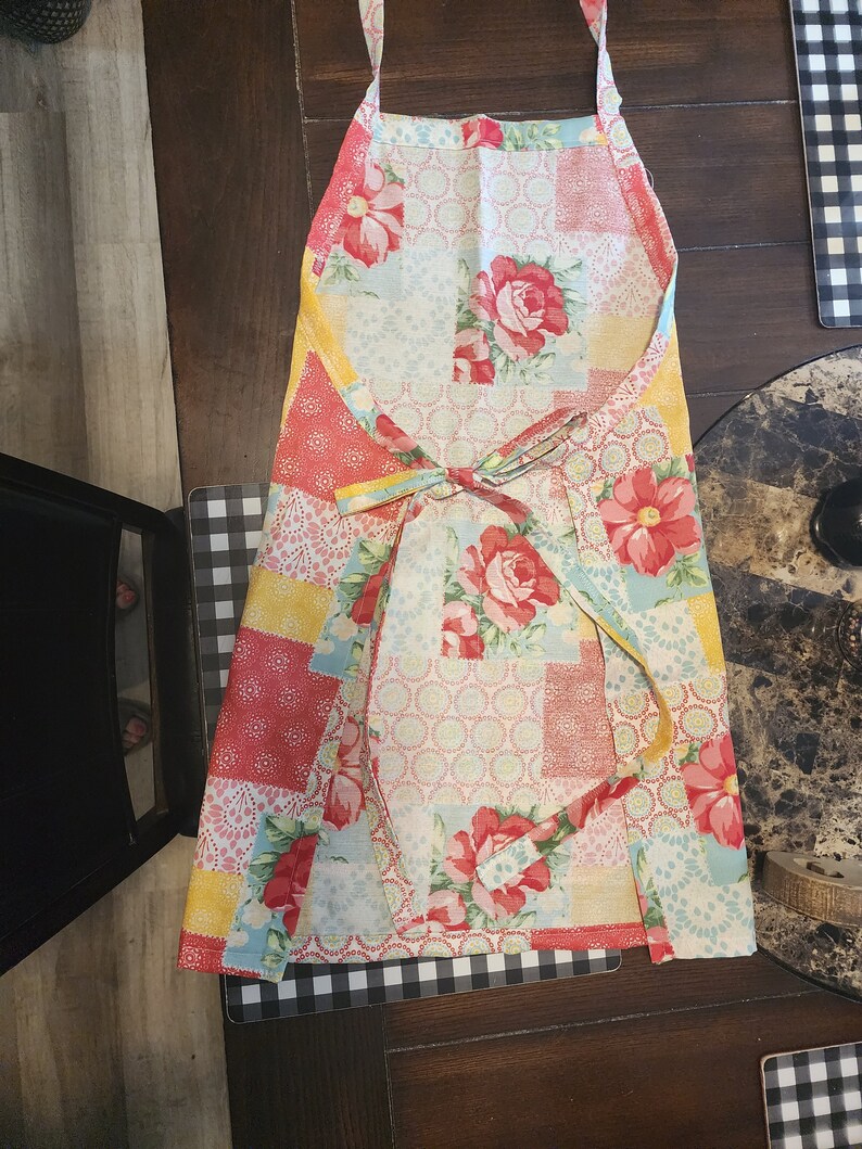 May include: A colourful patchwork apron with a floral pattern in shades of red, yellow, blue, and white. The apron has adjustable straps and a tie at the waist.