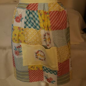 May include: A patchwork apron with a yellow waistband and pocket. The apron features a variety of patterned squares in yellow, red, blue, and white, with floral and striped designs. The pocket has a floral wreath design.
