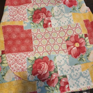 May include: A patchwork fabric with a floral pattern in shades of red, yellow, and blue. The fabric has a quilted texture and features a variety of floral designs, including roses and smaller flowers. The fabric is folded in half and the edges are visible.
