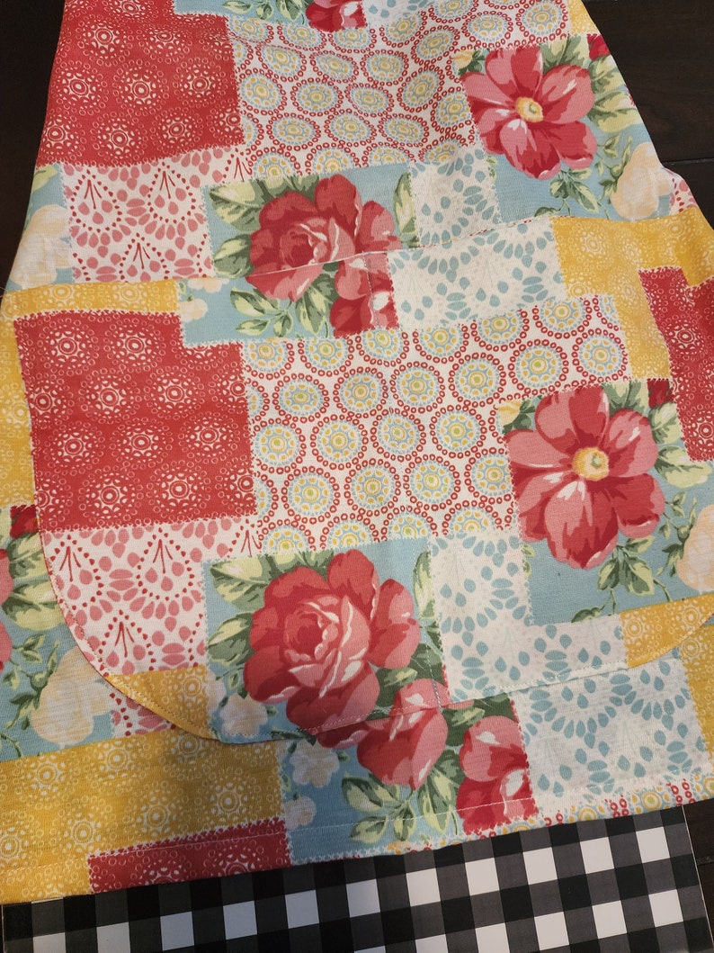 May include: A patchwork fabric with a floral pattern in shades of red, yellow, and blue. The fabric has a quilted texture and features a variety of floral designs, including roses and smaller flowers. The fabric is folded in half and the edges are visible.