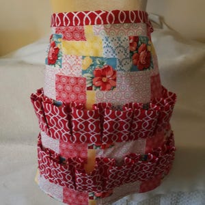 May include: A red and white patchwork apron with a ruffled bottom and multiple pockets. The apron has a floral print with red, yellow, blue, and white flowers.