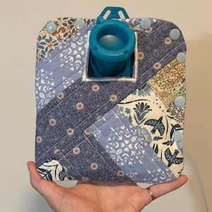 May include: A quilted water bottle cosy with a blue plastic spout and handle. The cosy is made of fabric with a patchwork design in shades of blue, cream, and green. Light blue buttons secure the cosy. A white cord extends from the bottom.