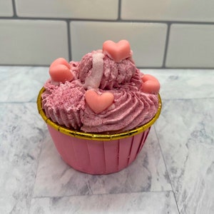May include: A pink cupcake with pink frosting and heart-shaped candies. The cupcake is in a pink paper liner with a gold rim.