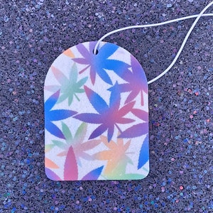 May include: A white car air freshener with a colorful ombre design of marijuana leaves. The leaves are in shades of pink, blue, green, and yellow.