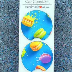 May include: Two car coasters with a blue background and a pattern of colorful macarons. The coasters have the text "Car Coasters" and "Handmade with love" on a white card.
