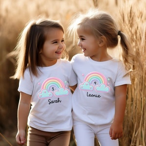 May include: Two girls wearing white t-shirts with rainbow and cloud designs. The shirts have the numbers 3 and 4 and the names Sarah and Leonie on them.