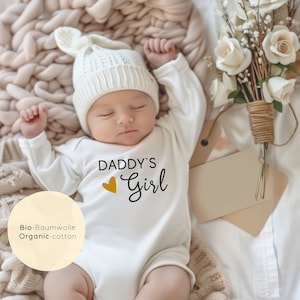 Pode incluir: Um macacão branco para bebê com o texto "Daddy's Girl" em letras pretas e um coração dourado. O macacão é feito de algodão orgânico.