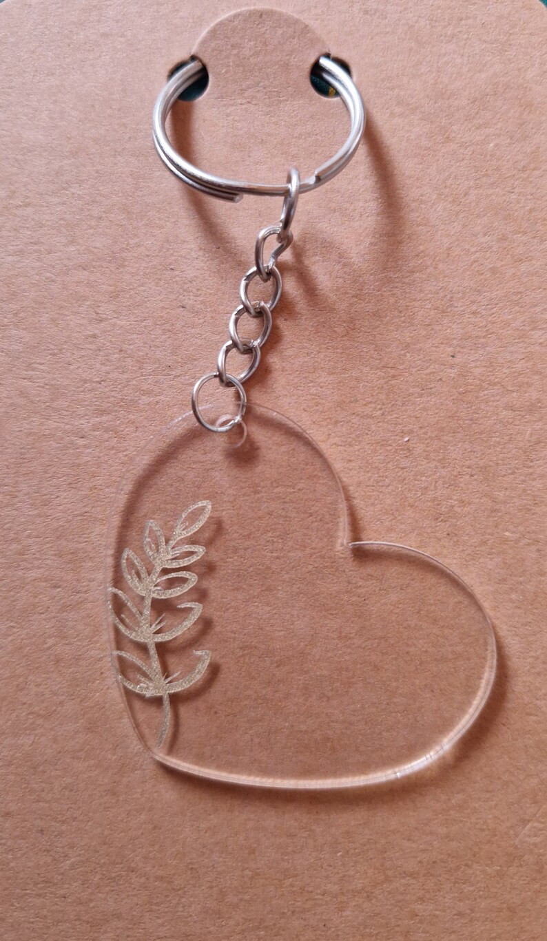 May include: A clear heart-shaped keyring with a silver chain and ring. A gold leaf design is etched into the heart.