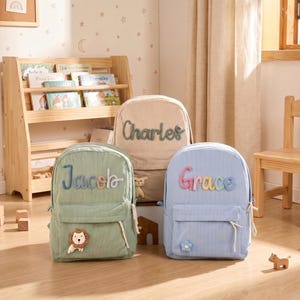 May include: Three corduroy backpacks in sage green, beige, and light blue. The sage green bag features the name Jacob and a lion patch. The beige bag displays the name Charles. The light blue bag shows the name Grace and a blue flower patch.