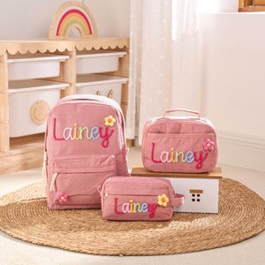 May include: Pink corduroy backpack, lunch bag, and pencil case set. Each item features the name "Lainey" in colorful embroidered letters and a flower accent. The set is designed for children and sits on a woven mat.