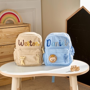 May include: Two corduroy backpacks, one beige and one light blue, sit on a white table. The beige backpack has the name "Weston" embroidered in brown and yellow, and a small animal applique. The blue backpack has "Daniel" embroidered in blue, and a lion applique.