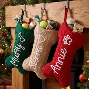 May include: Three cable-knit Christmas stockings in green, beige, and red hanging from a wooden mantel. The green stocking features the name Muffy and a bone icon, the beige one says Sierra with a heart, and the red one says Annie with a paw print.