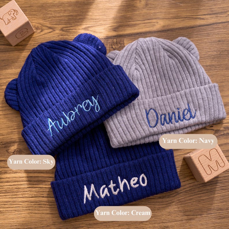 May include: Three personalized knit beanies in navy blue and gray, each with embroidered names. The navy beanies have sky and cream yarn colors, while the gray beanie has navy yarn. The hats have small ear-like details on top.