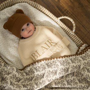 May include: A baby in a brown bear-eared knitted hat and cream sweater embroidered with the name "BLAIR" in gold. The baby lies in a woven basket, covered by a floral-patterned blanket.