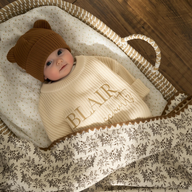 May include: A baby in a brown bear-eared knitted hat and cream sweater embroidered with the name "BLAIR" in gold. The baby lies in a woven basket, covered by a floral-patterned blanket.